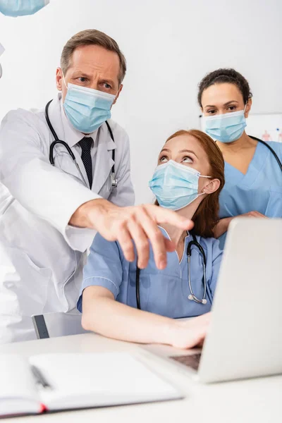 African american nurse standing near doctor pointing with finger at laptop near colleagues on blurred foreground — Stock Photo