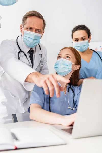 Enfermera afroamericana de pie cerca del médico señalando con el dedo a la computadora portátil cerca de colegas en primer plano borroso - foto de stock