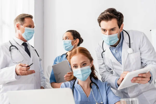 Медсестра в медичній масці дивиться на ноутбук біля мультикультурних колег в лікарні на розмитому передньому плані — стокове фото