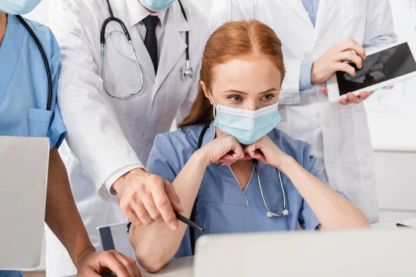 Руда медсестра в медичній масці дивиться на ноутбук біля мультикультурних колег, стоячи позаду на розмитому передньому плані — стокове фото