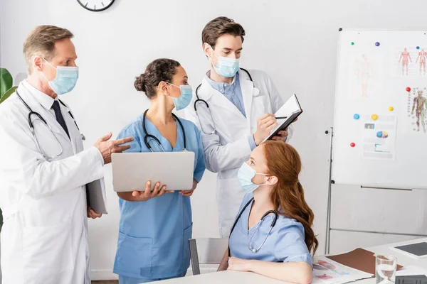 African american nurse with laptop looking at notebook while standing near colleagues in hospital — Stock Photo