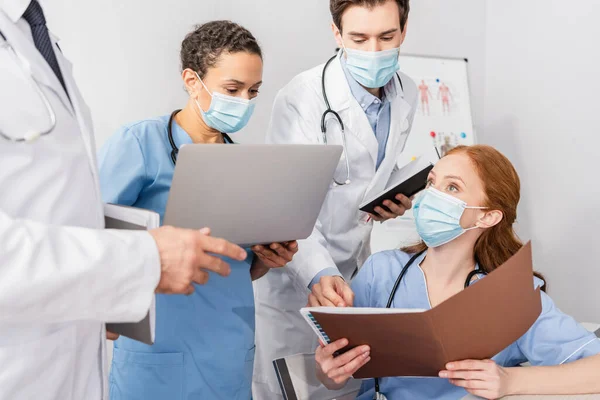 Krankenschwester mit medizinischer Maske hält Papiermappe in der Hand, während sie neben multiethnischen Kollegen im Krankenhaus sitzt — Stockfoto