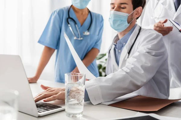 Arzt in medizinischer Maske mit Papiertippen auf Laptop neben multiethnischen Kollegen im verschwommenen Vordergrund im Krankenhaus — Stockfoto