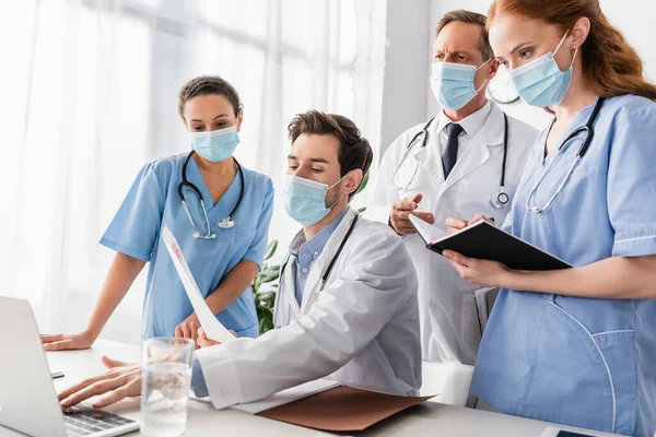 Colegas multiétnicos en máscaras médicas de pie cerca del médico escribiendo en el ordenador portátil en el lugar de trabajo en el hospital - foto de stock