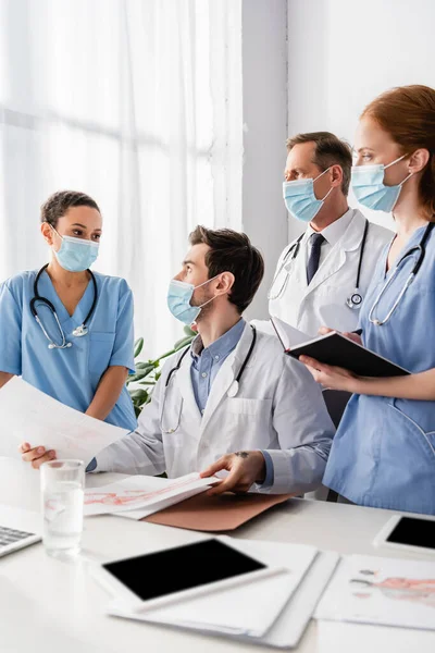 Krankenhauspersonal in medizinischen Masken betrachtet eine afrikanisch-amerikanische Krankenschwester, die in der Nähe ihres Arbeitsplatzes im verschwommenen Vordergrund steht — Stockfoto