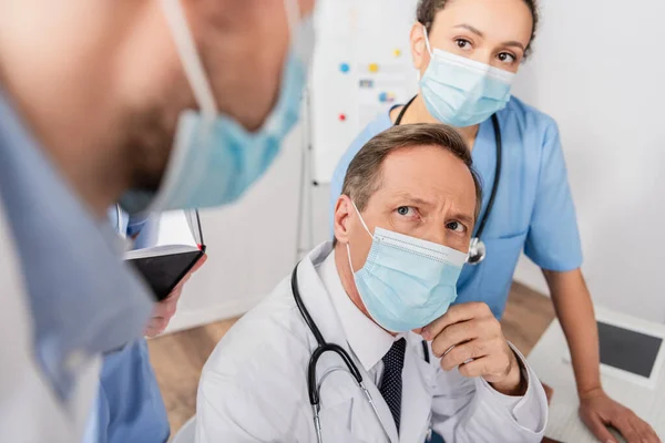 Multicultural doctor and nurse in medical masks looking at colleague on blurred foreground in hospital — Stock Photo