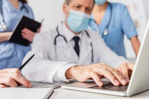 Close up view of doctor in medical mask typing on laptop near colleagues on blurred background — Stock Photo