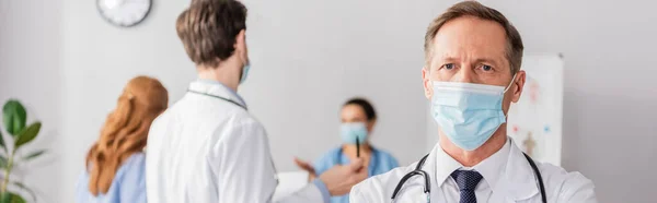 Doctor in medical mask looking at camera with blurred colleagues on background, banner — Stock Photo