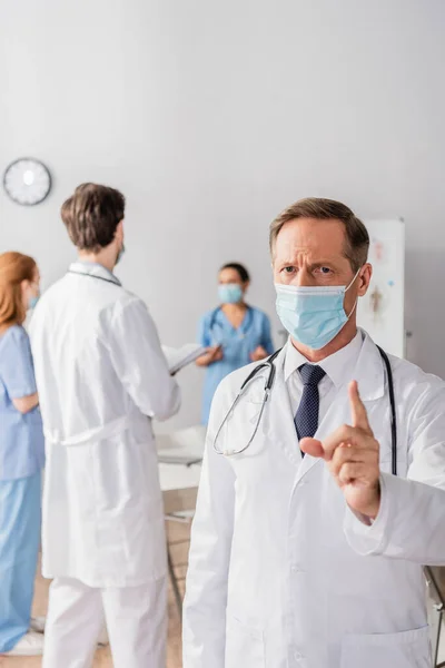 Doctor in medical mask with warning gesture looking at camera with blurred colleagues working on background — Stock Photo