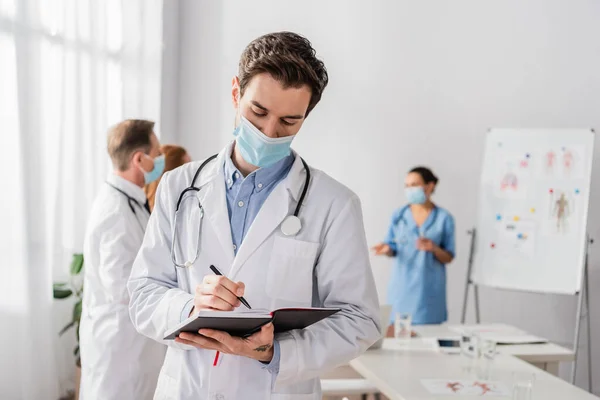 Doctor en escritura de máscaras médicas en cuaderno con colegas borrosos que trabajan en el fondo - foto de stock