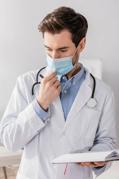 Vista frontale del medico che tocca la maschera medica mentre tiene il notebook e la penna in ospedale su sfondo sfocato — Foto stock