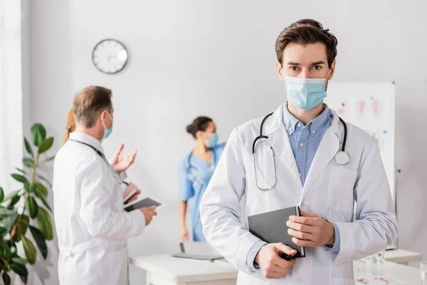 Arzt mit medizinischer Maske blickt in die Kamera, während er ein Notizbuch mit verschwommenen Kollegen hält, die am Hintergrund arbeiten — Stockfoto