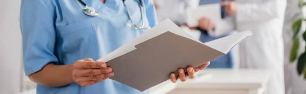 Cropped view of african american nurse holding paper folder with blurred colleagues on background, banner — Stock Photo