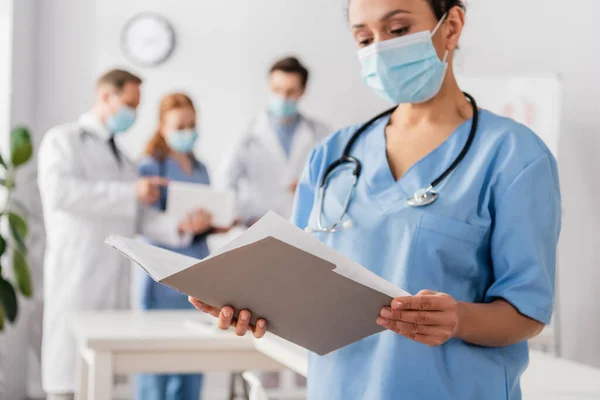 African american nurse looking at paper folder with blurred colleagues on background — Stock Photo