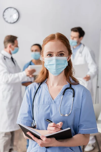 Vista frontale di infermiera rossa in maschera medica guardando la fotocamera con colleghi multiculturali offuscati sullo sfondo — Foto stock