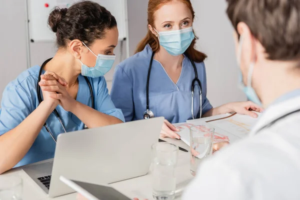 Multicultural hospital staff sitting at workplace with devices, papers and glasses of water on blurred foreground — Stock Photo