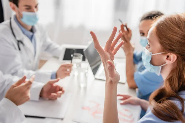 Enfermeira ruiva em gestos máscara médica enquanto conversa com colegas multiétnicos no local de trabalho em fundo turvo — Fotografia de Stock