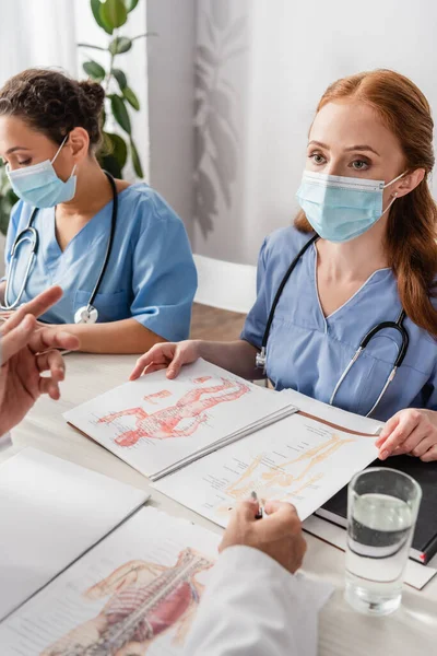 Doctor gesturing while sitting near multicultural colleagues at workplace with pictures in hospital — Stock Photo