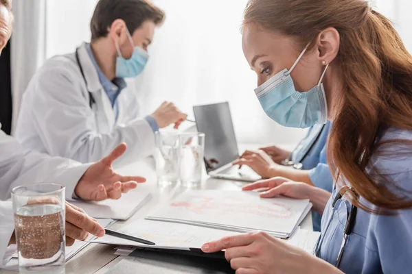 Infirmière rousse regardant dossier en papier avec des photos tout en étant assis près du médecin pointant avec un stylo près de collègues sur fond flou — Photo de stock