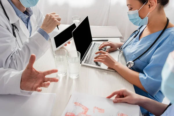 Африканская американская медсестра печатает на ноутбуке рядом с коллегами, работающими на рабочем месте с планшетом, бумагами и стаканами воды на размытом переднем плане — стоковое фото