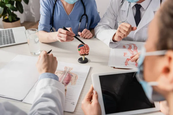 Infirmière pointant avec un stylo vers le modèle anatomique du cerveau près de collègues multiethniques sur le lieu de travail avec des images et des dispositifs au premier plan flou — Photo de stock