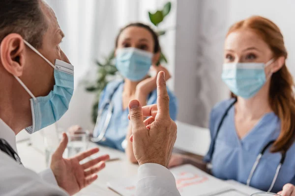 Médico em máscara médica apontando com o dedo enquanto sentado no local de trabalho com colegas multiétnicos desfocados em segundo plano — Fotografia de Stock