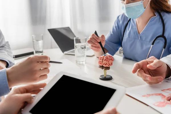 Обрезанный вид медсестры с цифровым планшетом рядом с коллегами на рабочем месте с ноутбуком и анатомической моделью мозга в больнице — стоковое фото