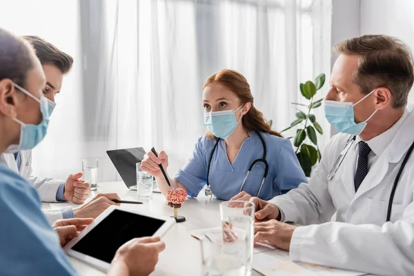 Enfermeras y médicos multiculturales que trabajan sentados en el lugar de trabajo con dispositivos, papeles y vasos de agua en un primer plano borroso - foto de stock