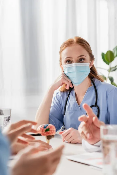 Redhead nurse in medical mask sitting at workplace with brain anatomical model near colleagues on blurred foreground — Stock Photo