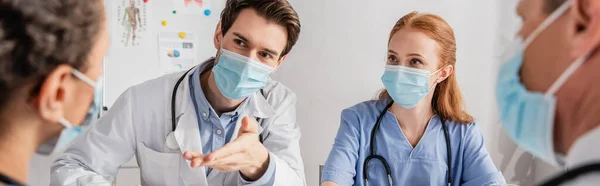 Доктор в медичній масці, сидячи поруч з колегами і вказуючи рукою на афроамериканську медсестру в лікарні на розмитому передньому плані, банер — стокове фото