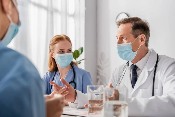 Médico em máscara médica conversando com colegas enquanto sentado no local de trabalho com copos borrados de água em primeiro plano — Fotografia de Stock