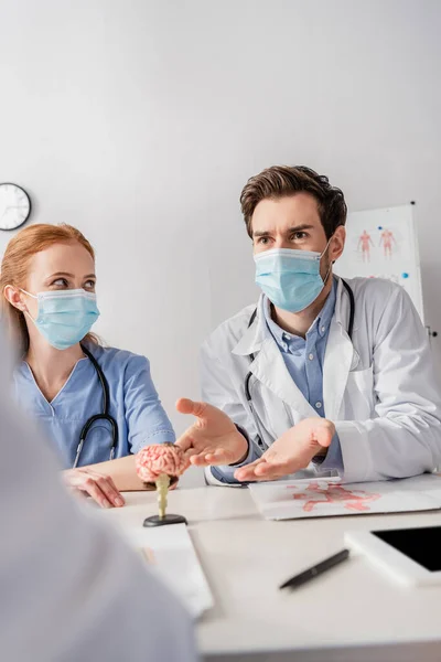 Médico en máscara médica hablando con colegas mientras está sentado en el lugar de trabajo con papeles y modelo anatómico del cerebro en primer plano borroso - foto de stock