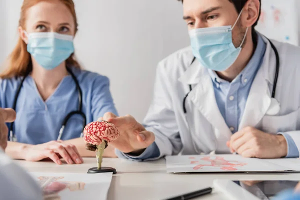 Arzt in medizinischer Maske berührt anatomisches Modell des Gehirns, während er am Arbeitsplatz in der Nähe von Kollegen im verschwommenen Vordergrund sitzt — Stockfoto