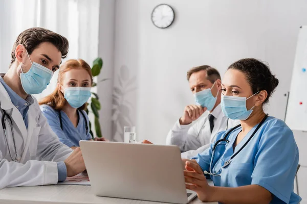 Multicultural hospital staff in medical masks looking at laptop while sitting at workplace in hospital on blurred background — Stock Photo