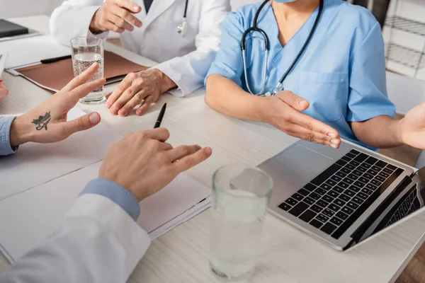 Ausgeschnittene Ansicht einer afrikanisch-amerikanischen Krankenschwester, die mit der Hand auf einen Laptop zeigt, in der Nähe von Kollegen am Arbeitsplatz mit Papieren im Krankenhaus — Stockfoto