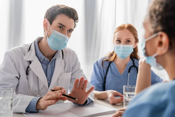 Arzt in medizinischer Maske sitzt neben Krankenschwester und spricht mit afrikanisch-amerikanischem Kollegen am Arbeitsplatz auf verschwommenem Vordergrund — Stockfoto