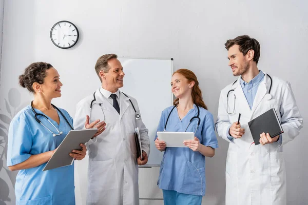 Happy multicultural doctors and nurses looking at each other while standing together with flipchart and wall clock on background — Stock Photo