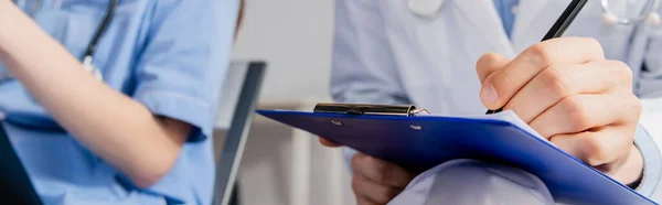 Cropped view of doctor writing on clipboard near colleague during meeting in hospital on blurred background, banner — Stock Photo