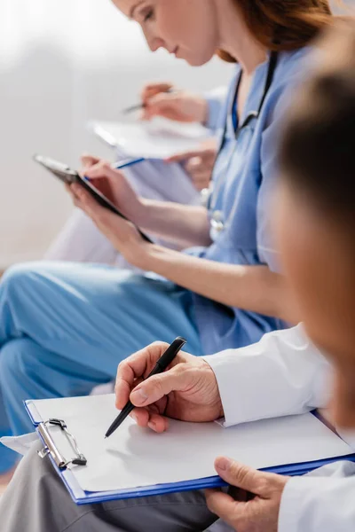 Doctor writing on clipboard near multicultural colleagues during meeting in hospital on blurred foreground — Stock Photo