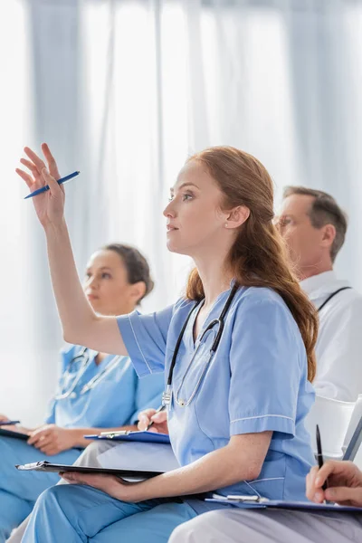 Rothaarige Krankenschwester mit erhobener Hand sitzt neben multikulturellen Kollegen während eines Treffens im Krankenhaus auf verschwommenem Hintergrund — Stockfoto