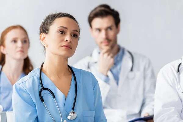 African american nurse looking away during meeting with blurred colleagues on background — Stock Photo