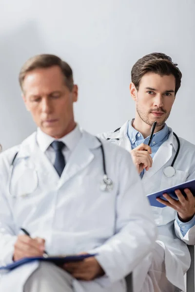 Médico reflexivo con lápiz y portapapeles mirando a la cámara durante la reunión con un colega borroso en primer plano en el hospital - foto de stock