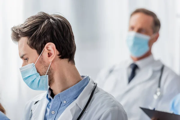 Doctor in medical mask during meeting in hospital with blurred colleague on background — Stock Photo