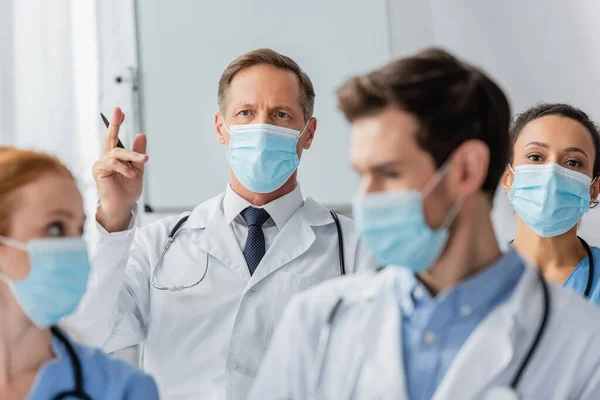 Doctor with raised hand near african american nurse during meeting with blurred colleagues on foreground in hospital — Stock Photo