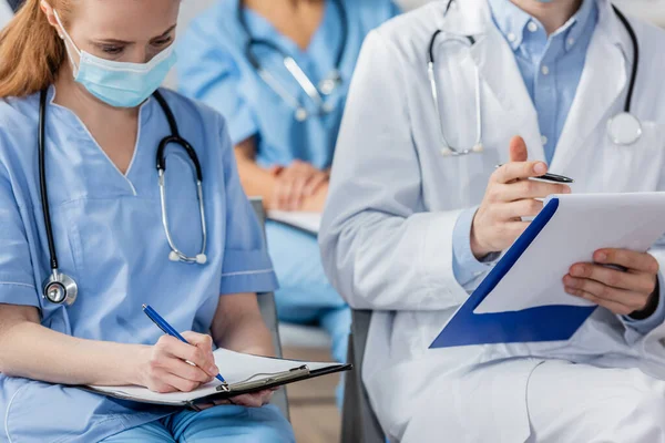 Krankenschwester schreibt auf Klemmbrett, während sie in der Nähe des Arztes sitzt, während sie sich mit verschwommenen Kollegen im Krankenhaus trifft — Stockfoto