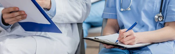 Nurse writing on clipboard while sitting near doctor during meeting in hospital on blurred background, banner — Stock Photo