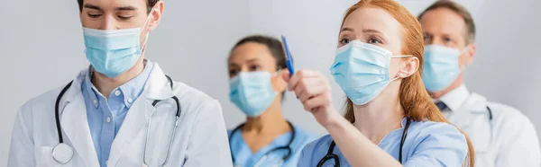 Enfermera pelirroja en máscara médica señalando con pluma durante la reunión con colegas multiétnicos borrosas en el fondo, pancarta - foto de stock