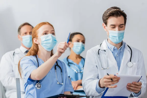 Рыжая медсестра указывает ручкой, сидя рядом с врачом во время встречи с размытыми коллегами на заднем плане в больнице — стоковое фото