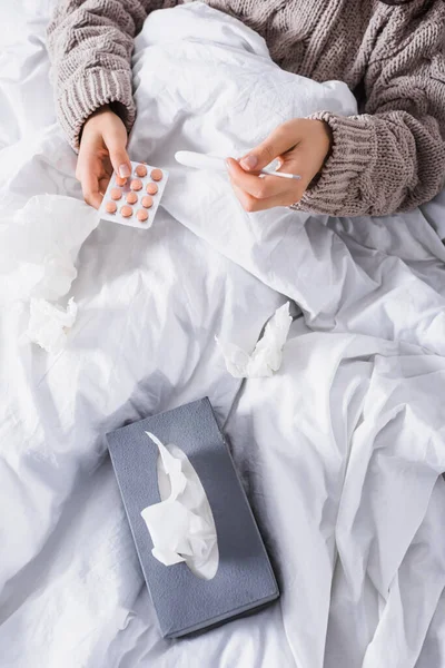 Vista ritagliata di giovane donna malata con tessuto, pillole e termometro a letto — Foto stock