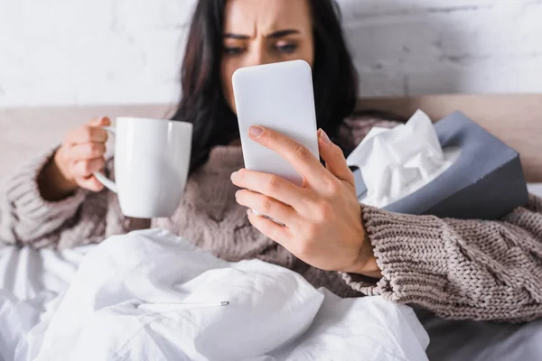 Malata giovane bruna donna con bevanda calda e tessuto che tiene smartphone a letto, sfondo sfocato — Foto stock